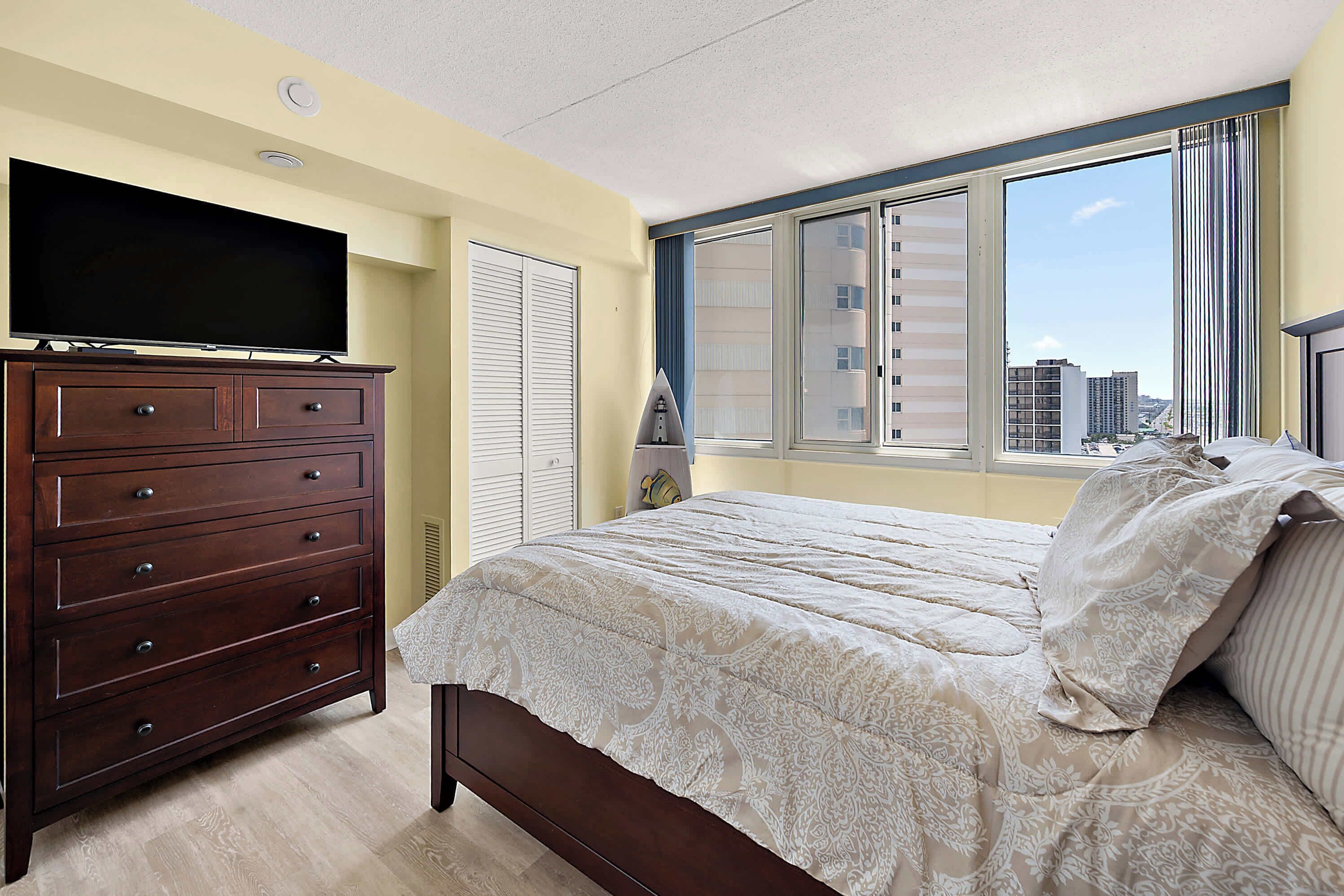 Ein Schlafzimmer mit Bett, Kommode, Fernseher und einem Fenster mit Blick auf die Stadt.
