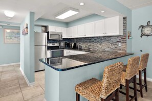 A kitchen with white cabinets, a stainless steel refrigerator, a microwave, and a stove. There is a bar area with woven chairs. - Two Bedroom Beachfront Condo with Great Amenities (Ocean City)
