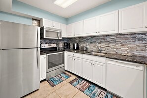 A modern kitchen with white cabinets, stainless steel appliances, a stone backsplash, and a doormat with 'HOME' and 'KITCHEN' on it. - Two Bedroom Beachfront Condo with Great Amenities (Ocean City)