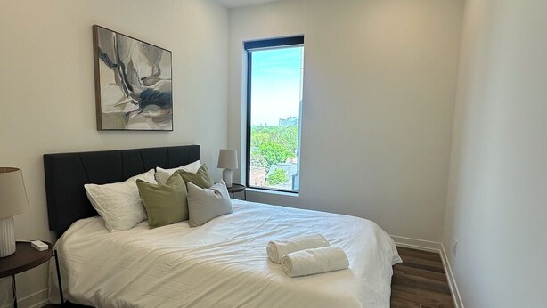 A bedroom with a bed, a nightstand, a lamp, a painting, and a window with a view. - Bright, Spacious 3 Bedroom Penthouse in Vibrant Leslieville in Downtown Toronto (Toronto)