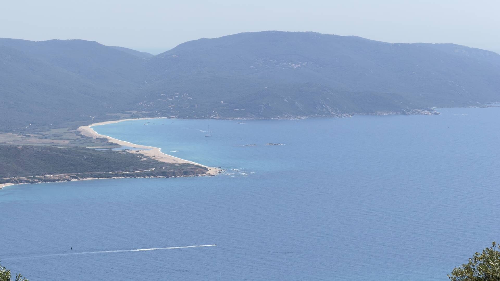 A coastal landscape with a beach, clear blue water, and distant mountains.