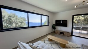 Ein modernes Wohnzimmer mit großem Fenster, Flachbildfernseher, einer Bank und einem Essbereich mit Stühlen.