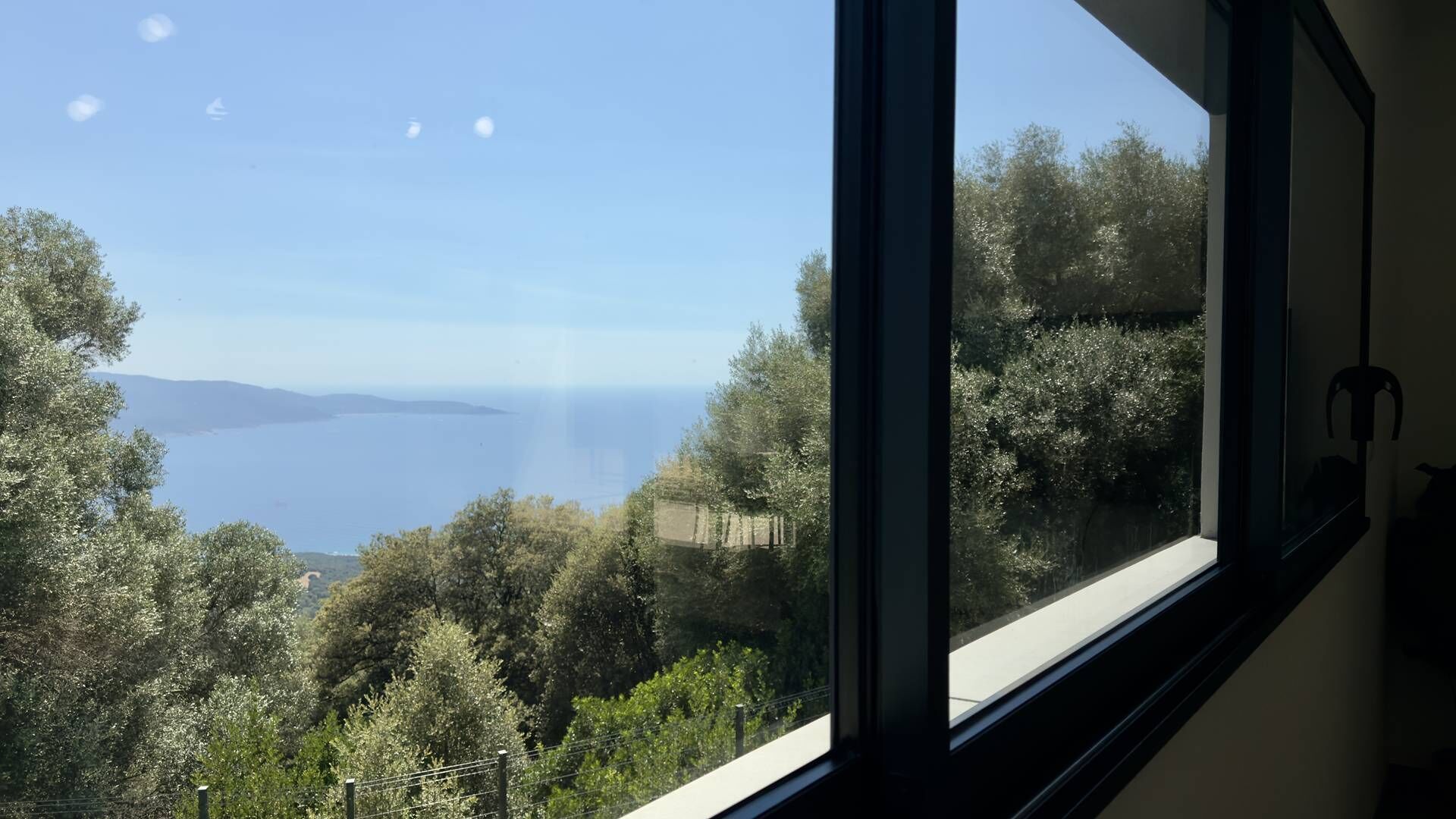 Une pièce dotée d’une grande fenêtre donnant sur un paysage côtier, avec des arbres et une vue sur la mer.