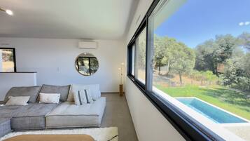 Een moderne woonkamer met een groot raam, een ronde spiegel en uitzicht op een zwembad.