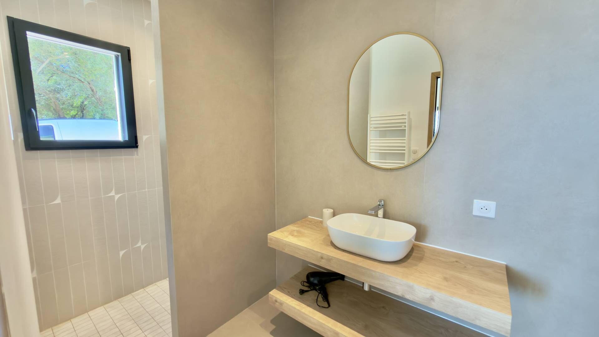A bathroom with a large mirror, a white sink, and a window with a view of trees.