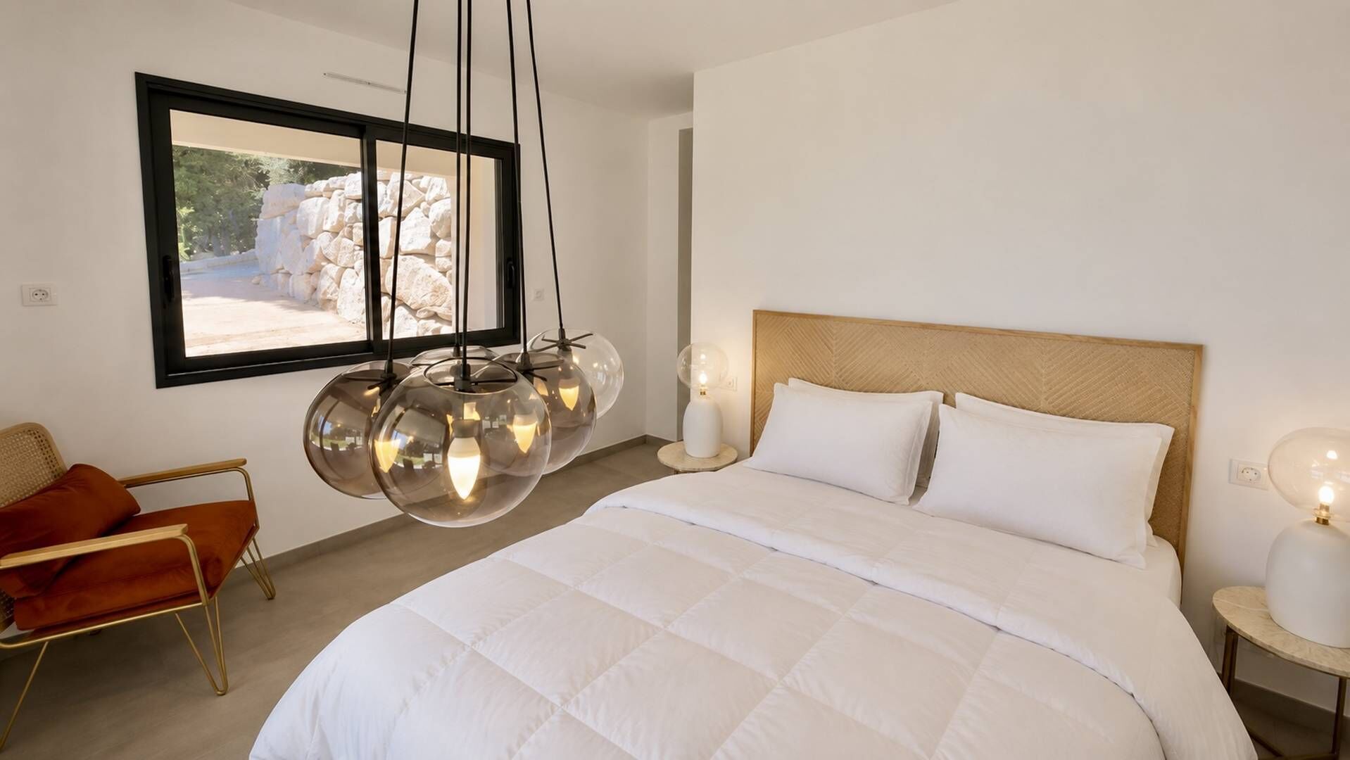 A bedroom with a large bed, a modern chandelier, a view of a stone wall, and a chair with cushions.