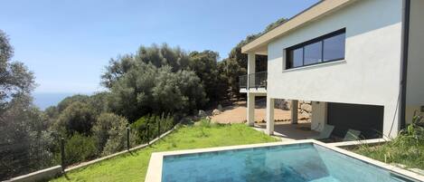 A modern two-story house with a swimming pool, surrounded by greenery and overlooking a hilly landscape.