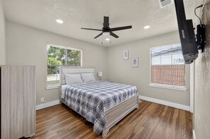 Una camera da letto con un letto, una ventola a soffitto, due finestre con tende e una TV a parete.