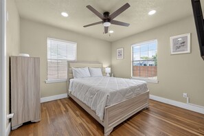 Ein Schlafzimmer mit einem Bett, einem Deckenventilator, zwei Fenstern mit Jalousien, einem Nachttisch mit Lampe und einem gerahmten Bild an der Wand.