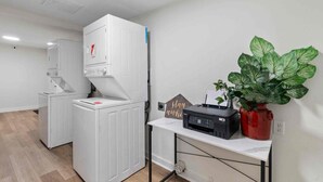 A laundry area with stacked washer and dryer units, a printer on a desk, and a potted plant. - Emerald Escape, a Central 4BR/5BA Home with Game Room, Large Yard & Walk to UCA! (Conway)