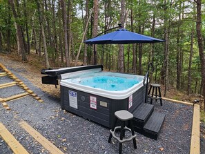 A hot tub with a safety sign, surrounded by a wooden deck and a forested area. - Bear Lodge - Hot tub & Bryce Mountain views! (Basye)