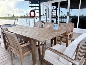 A wooden deck with a table and chairs, a lifebuoy, and a staircase leading to an upper deck.