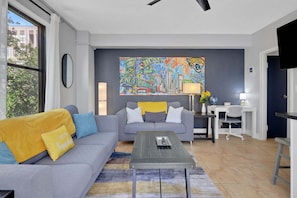 A modern living room with a grey sofa, a coffee table, and a colorful cityscape painting on the wall. - Downtown Jacksonville Retreat | Walk to Events & Stadium (Jacksonville)