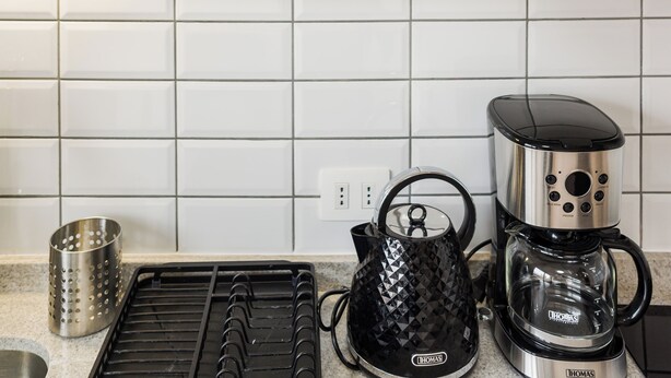 Et køkken med kaffemaskine, elkedel og en hylde med tallerkener og kopper.