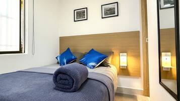 Quarto de hotel com uma cama, duas almofadas, um cobertor dobrado, uma mesinha de cabeceira com abajur e duas fotografias emolduradas na parede.