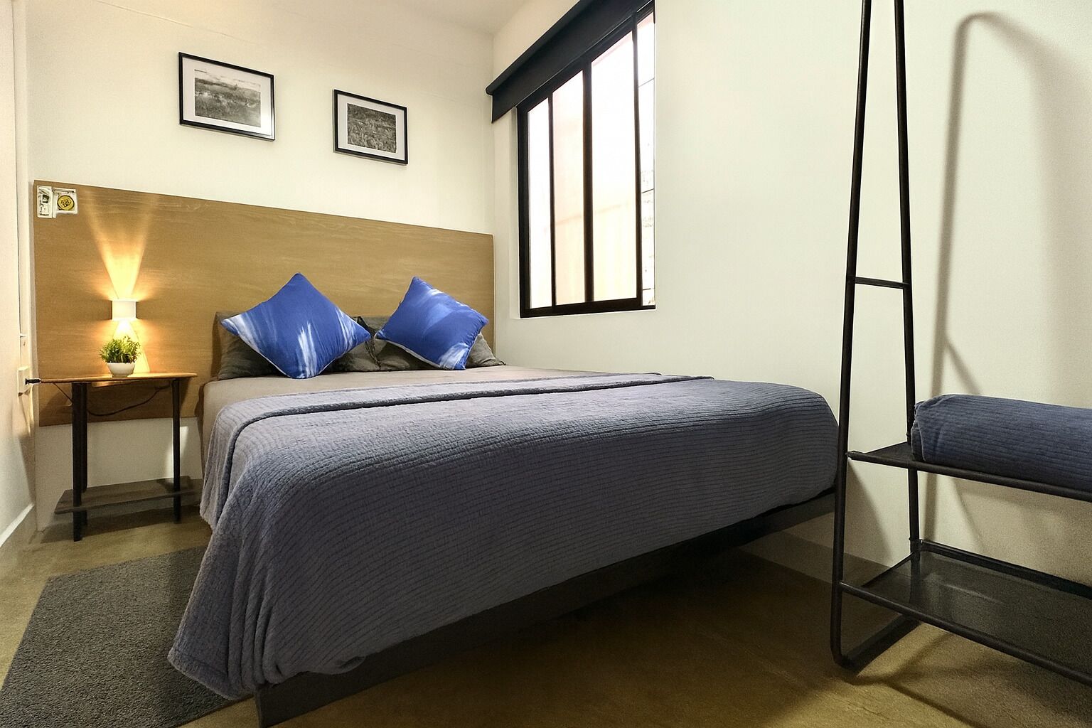 Un dormitorio con una cama, una mesita de noche con lámpara, dos cuadros enmarcados en la pared y una ventana.