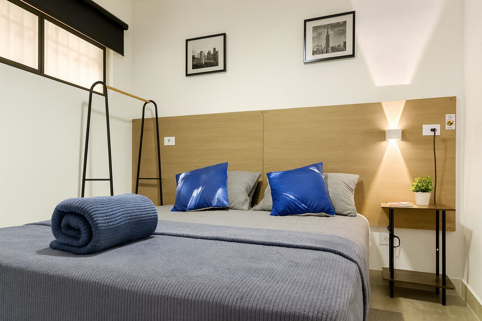 Habitación de hotel con cama, almohadas, mesita de noche con lámpara, una planta pequeña y dos cuadros en la pared.