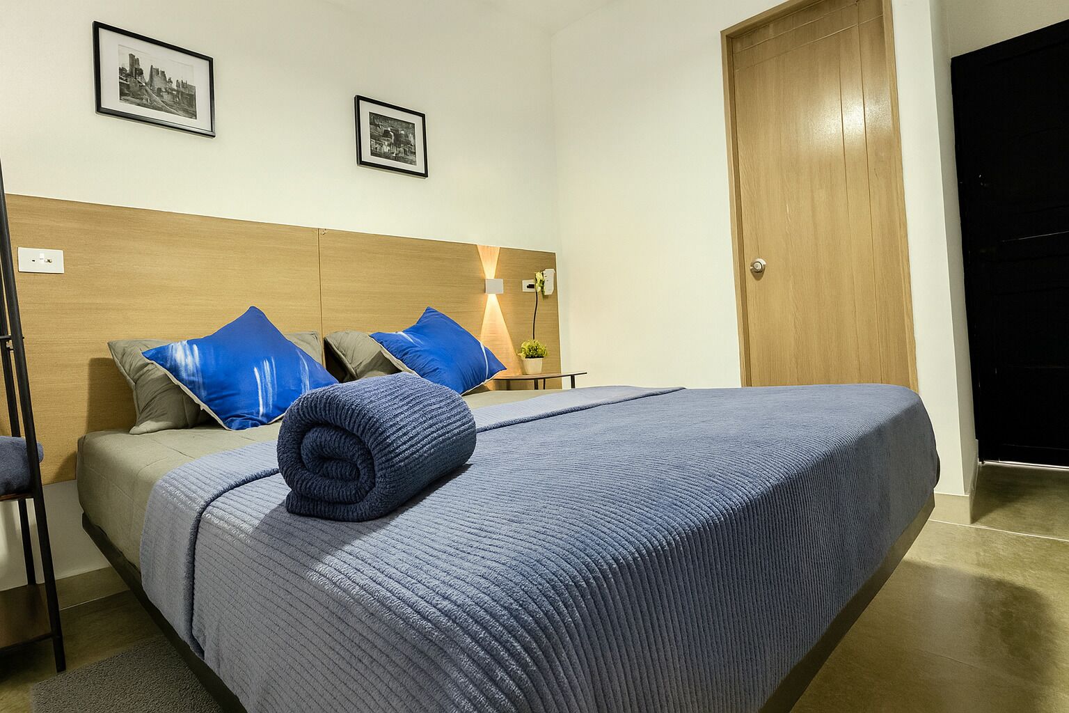 Una habitación de hotel con cama, almohadas, mesita de noche, lámpara y puerta.