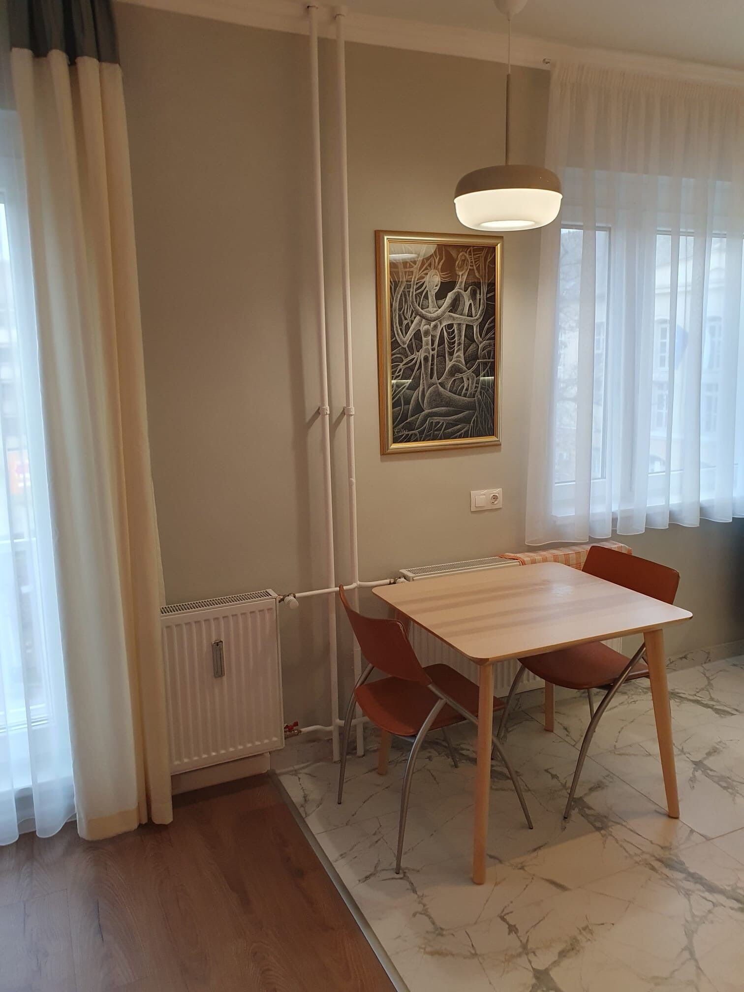 Un coin repas comprenant une table et des chaises, un radiateur, un luminaire au plafond et une fenêtre avec des rideaux.
