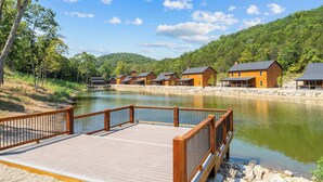Una terraza de madera con borde de piedra, un río tranquilo y varias cabañas de madera con techos inclinados.