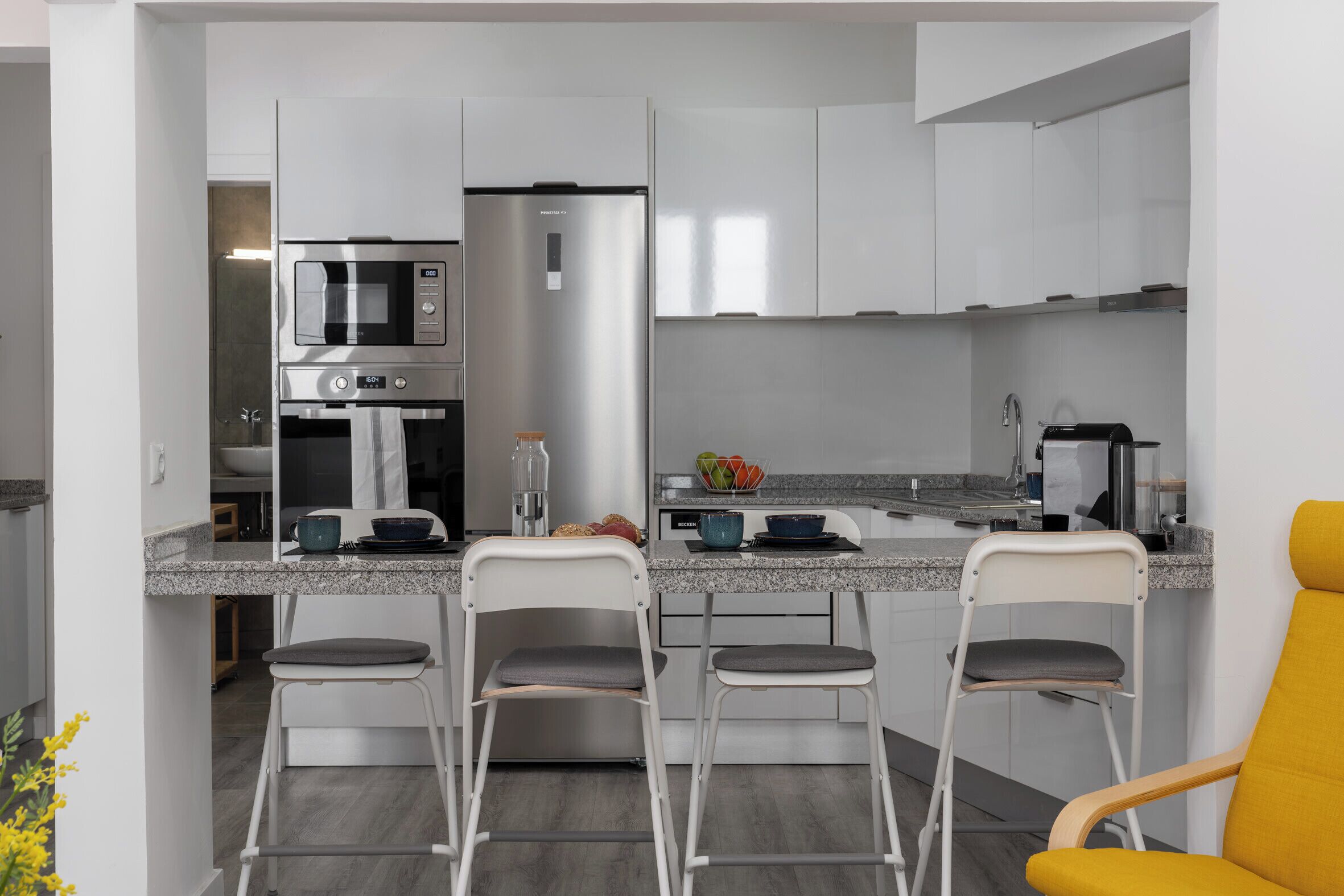 A modern kitchen with a built-in microwave, a stainless steel refrigerator, and a granite countertop. There are three bar stools and a yellow chair.