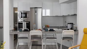 A modern kitchen with a built-in microwave, a stainless steel refrigerator, and a granite countertop. There are three bar stools and a yellow chair.