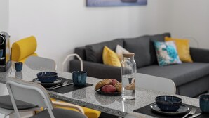 A modern living room with a dining area. There is a sofa with cushions, a coffee table with a glass bottle and pastries, and a chair with a cushion. - Funchal Oh My! - Cozy Stay with Terrace (Funchal)