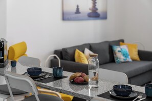 Ein modernes Wohnzimmer mit Essbereich. Es gibt ein Sofa mit Kissen, einen Couchtisch mit einer Glasflasche und Gebäck, sowie einen Sessel mit Kissen.