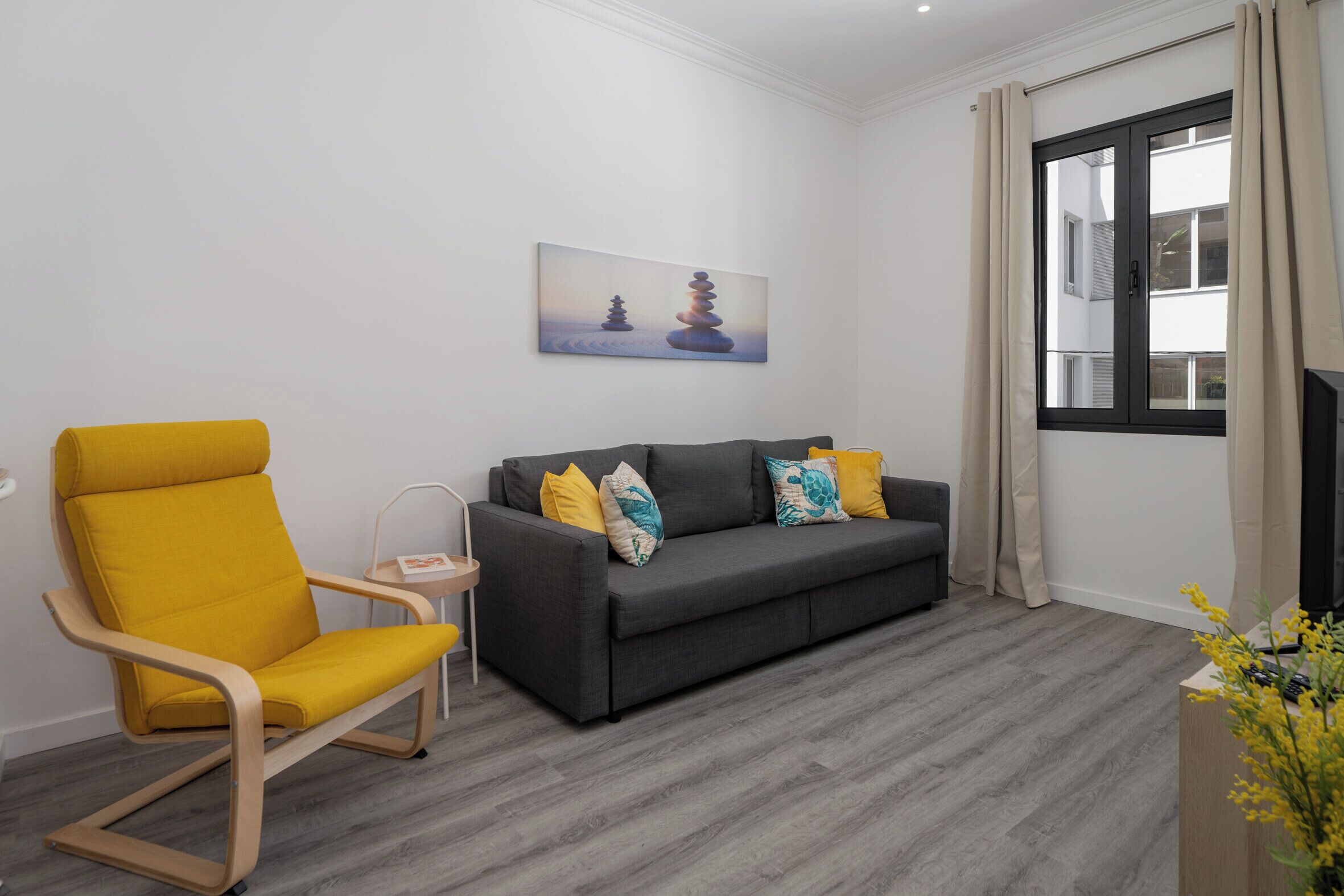 A living room with a grey sofa, yellow armchair, and a painting of stacked stones.