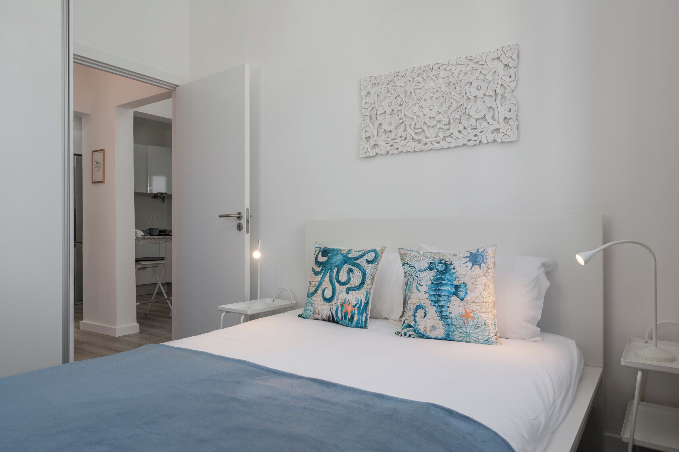 Un dormitorio con una cama, dos almohadones decorativos, una obra de arte en la pared, una lámpara de noche y vistas a una cocina.