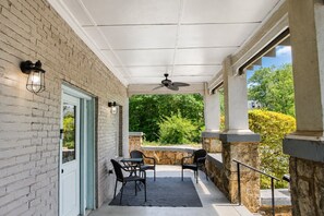 A covered patio with a brick wall, a door, two chairs, a table, a ceiling fan, and a view of greenery.