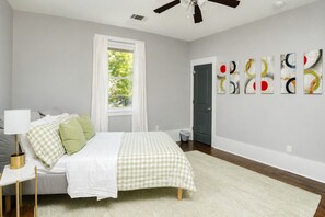 A bedroom with a bed, a bedside table, a lamp, a ceiling fan, a window with curtains, and abstract wall art.