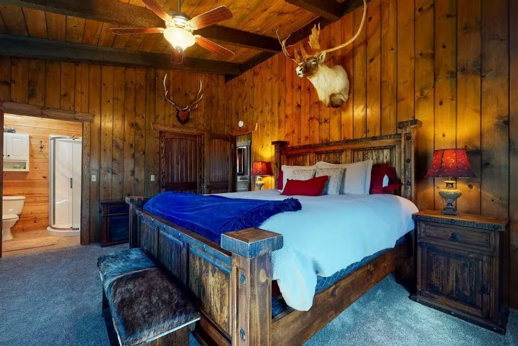 A wooden cabin bedroom with a bed, a ceiling fan, a mounted deer head, and a nightstand with a lamp.