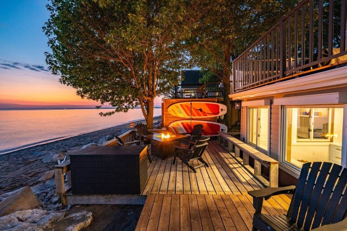 Une terrasse en bois avec des sièges d’extérieur et des planches de surf, donnant sur une plage au coucher du soleil.