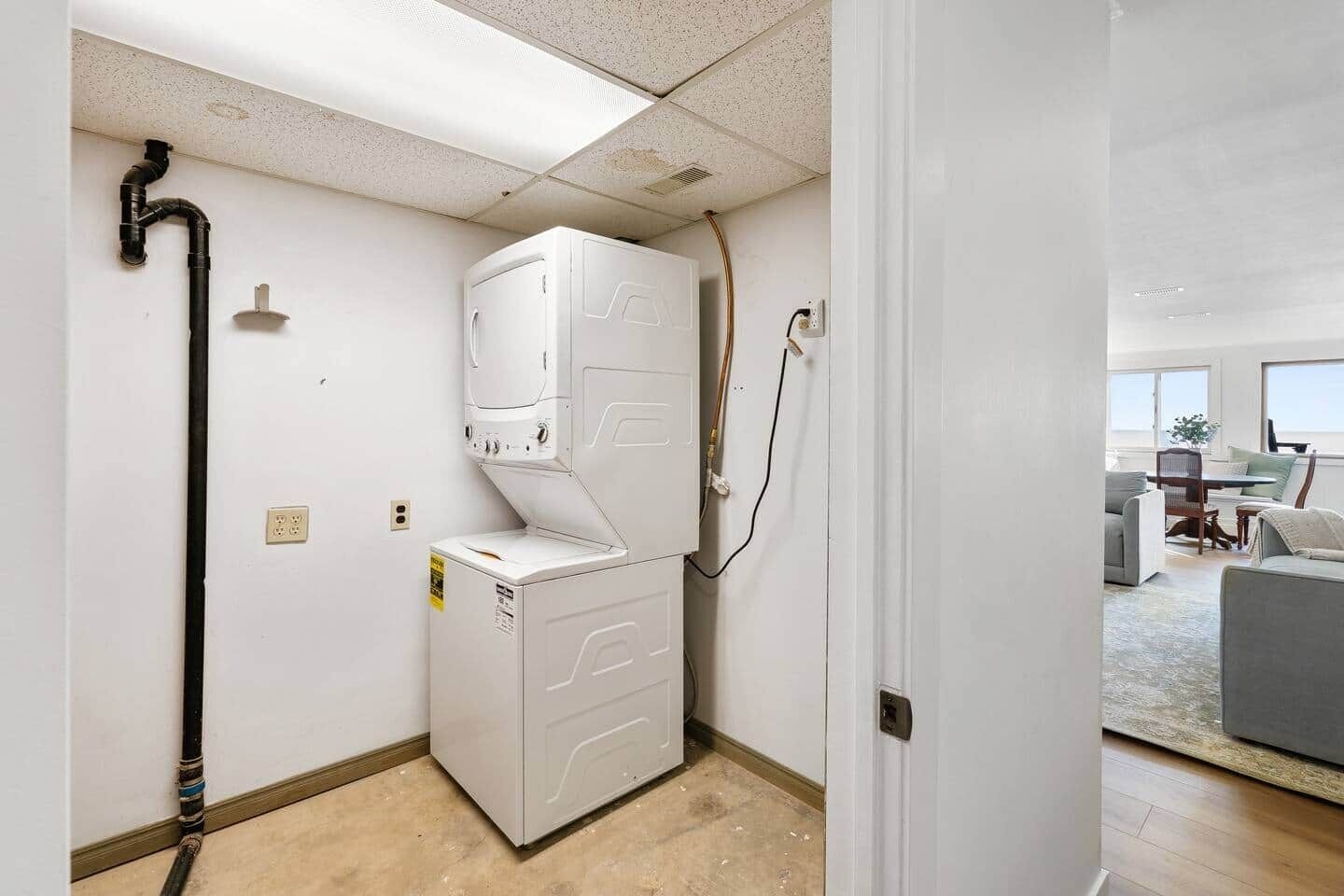 A compact laundry room with a washer and dryer, a visible water pipe, and a door leading to a living area with a sofa and a table.