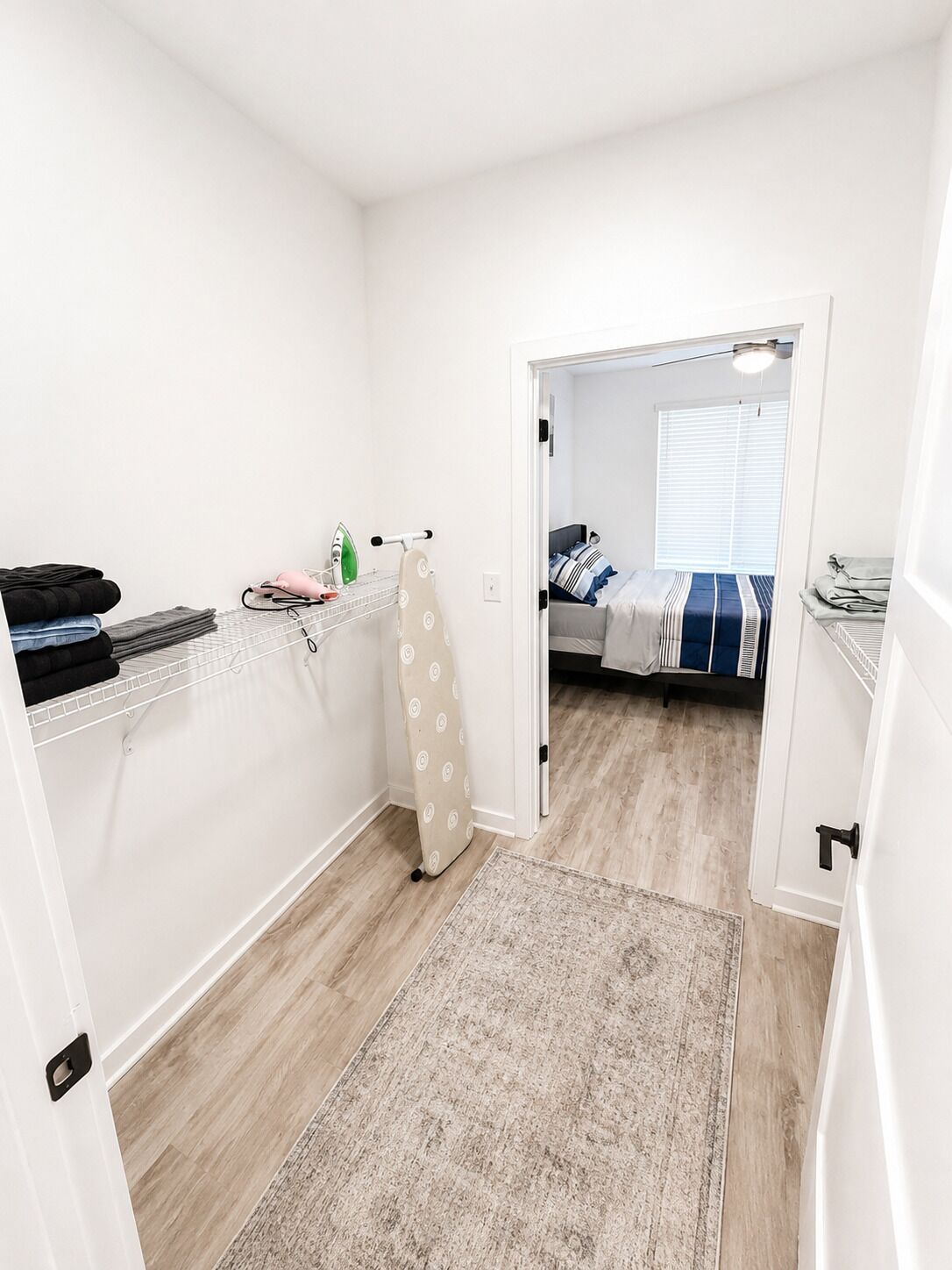 A compact bedroom with a bed, a small ironing station, and a door leading to another room.