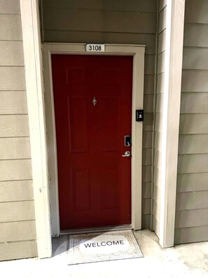 A red door with a number plate '3108' above it, a doorbell, and a 'WELCOME' mat at the entrance. - Resort-Style 1BR Pool, Gym & Movie Theater (Conroe)