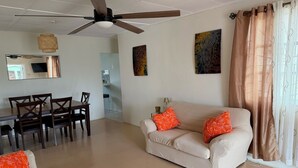 Ein Wohnzimmer mit Deckenventilator, einem Esstisch mit Stühlen, einer Couch mit orangefarbenen Kissen und Bildern an den Wänden.