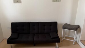 Ein schwarzes Sofa mit Metallbeinen, ein kleiner weißer Hocker und eine schlichte weiße Wand mit einer Türspiegelung.