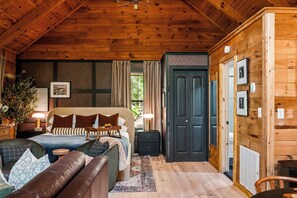 A cozy wooden cabin bedroom with a bed, bedside tables, a dresser, and a large window. - Studio cabin in the Smokies. Sauna, hot tub, fire pit, private deck, EV charger (Sevierville)