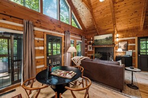 A cozy cabin interior with a fireplace, a dining area with a round table, and a comfortable seating arrangement. - Studio cabin in the Smokies. Sauna, hot tub, fire pit, private deck, EV charger (Sevierville)