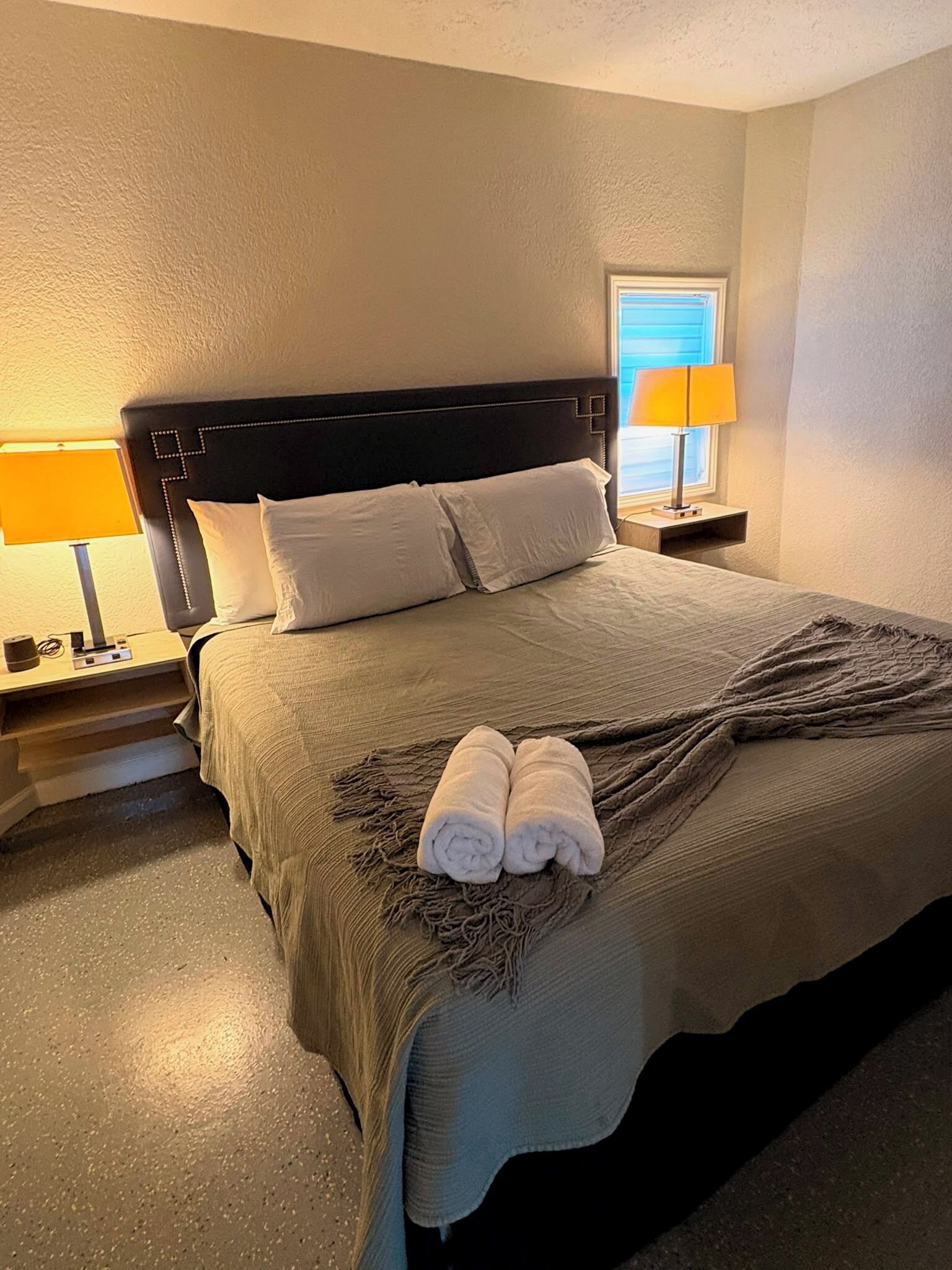 Una cama bien hecha con un cabecero oscuro, dos lámparas de noche y una ventana con persianas.