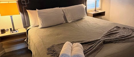 Una cama bien hecha con un cabecero oscuro, dos lámparas de noche y una ventana con persianas.
