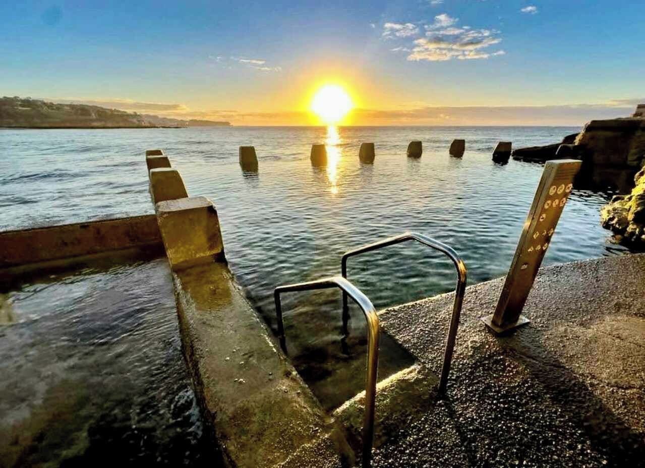 Um cais de concreto com grades de metal que se estende por águas calmas, com um pôr do sol ao fundo e céu claro.