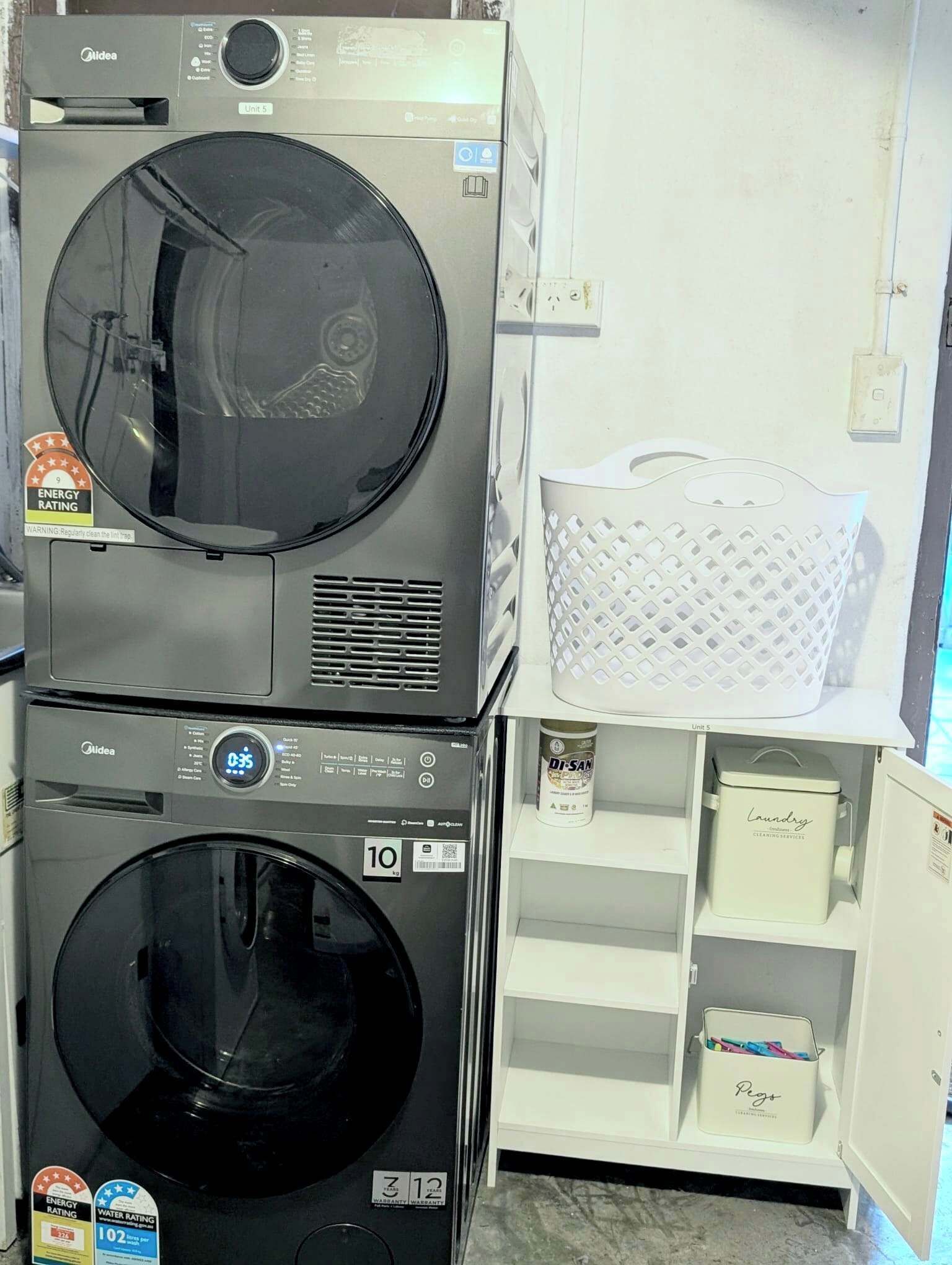 A laundry room with two stacked washing machines, a shelf with a basket, and a can of detergent.