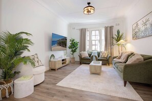 Ein modernes Wohnzimmer mit einer Couch, Sesseln, einem Couchtisch und einem Fernseher. Es befinden sich Pflanzen, ein Teppich und ein Gemälde an der Wand.