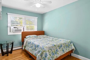 A bedroom with a bed, a window with blinds, a ceiling fan, and a small table.
