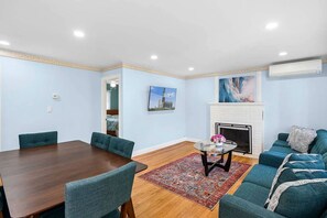 A room with a dining table, chairs, a sofa, a fireplace, and a painting on the wall. - Charming Hampton Bays Home Near Beaches (Hampton Bays)