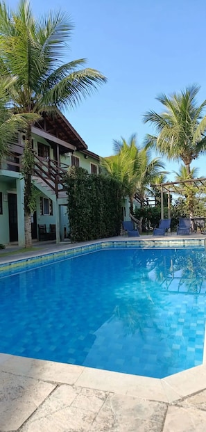 A swimming pool with lounge chairs, surrounded by palm trees and a building with a sloped roof. - Feet in the Sand / Beach House / Bertioga (Bertioga)