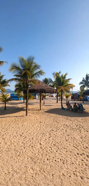 A sandy beach with palm trees and a thatched-roof structure. - Feet in the Sand / Beach House / Bertioga (Bertioga)